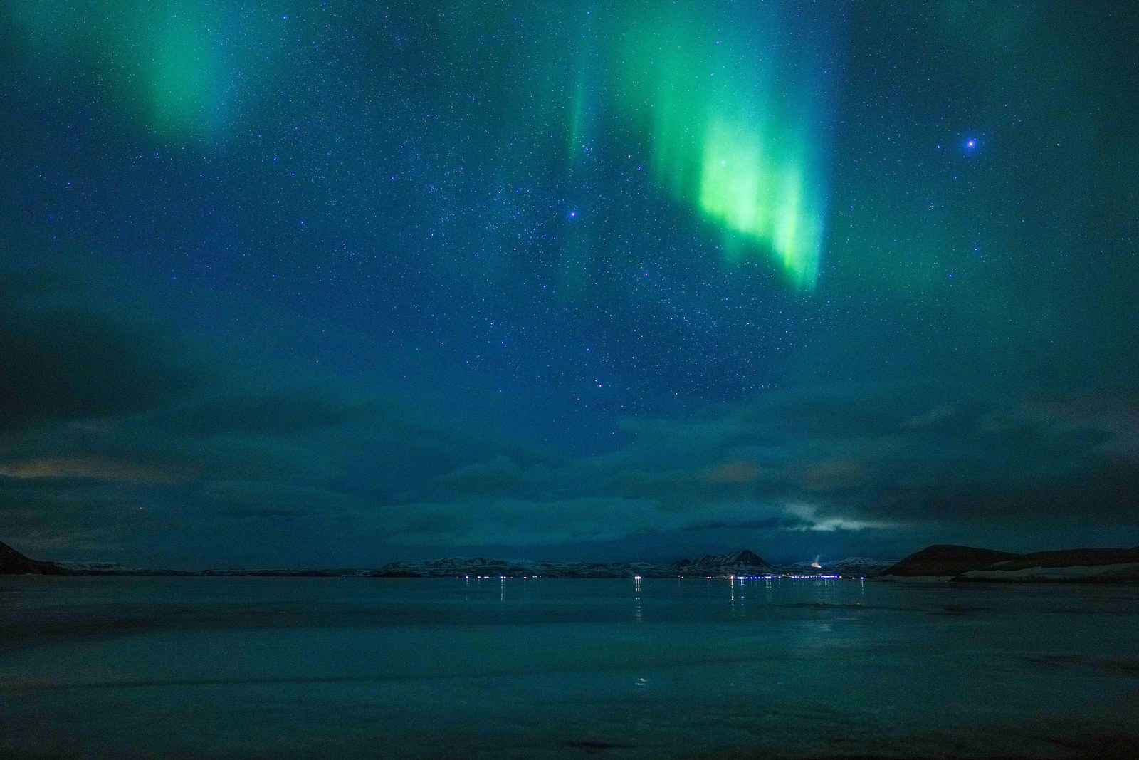 Green northern lights and stars above Lake Mývatn in Iceland, reflected in the dark water.
