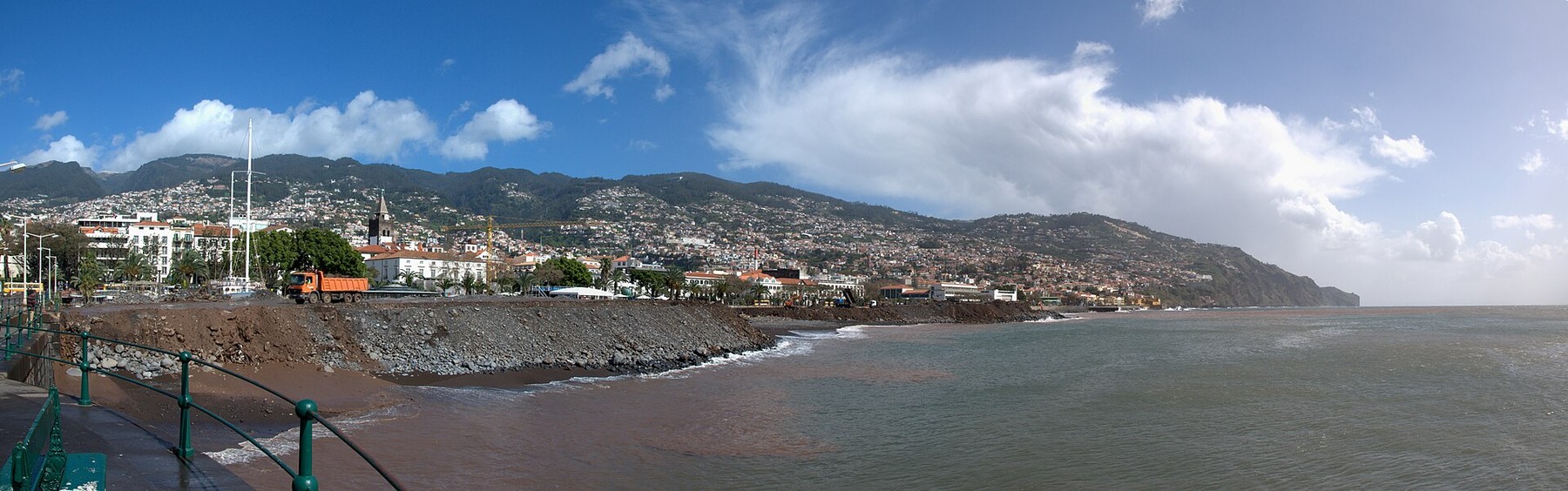 Funchal's waterfront and hillside city curve around a calm Atlantic bay beneath green mountains and a blue sky.