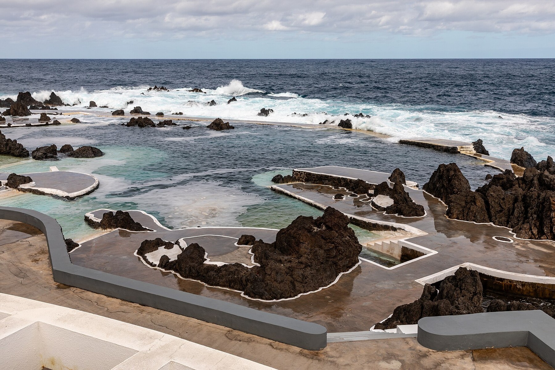 Natural seawater swimming pools edged by dark volcanic rock and pale walkways in Porto Moniz, with Atlantic waves beyond.